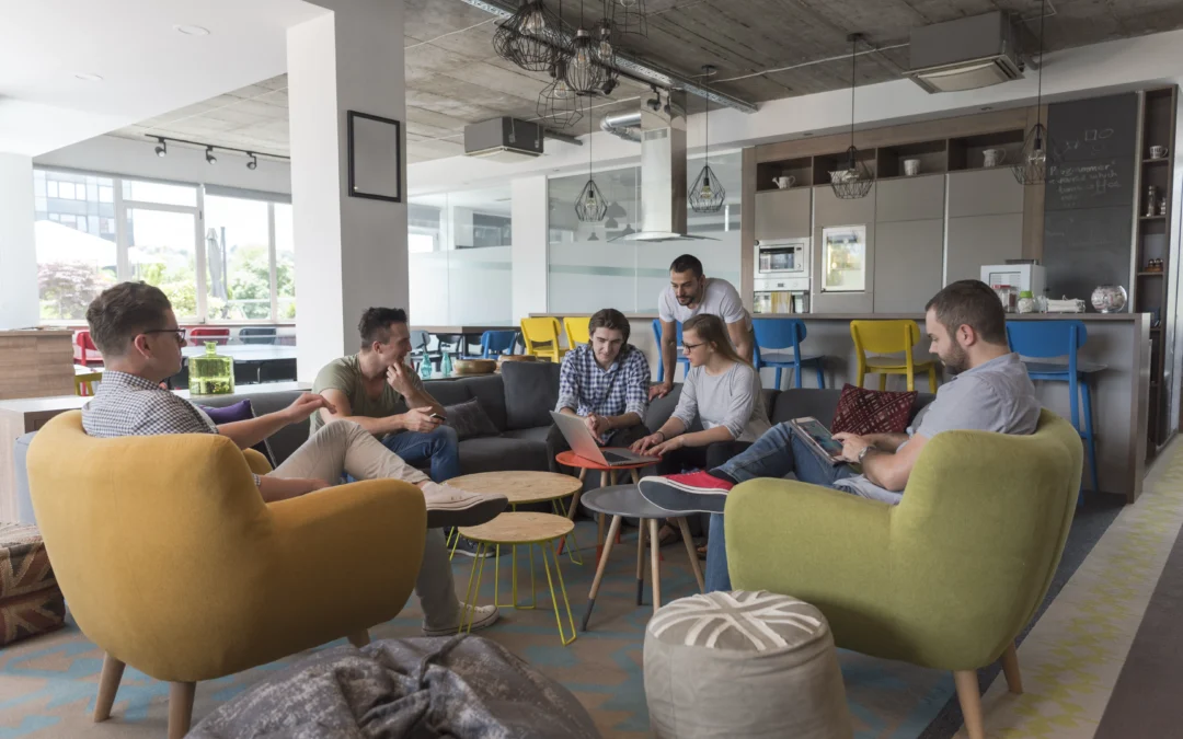 Group of coworkers seated in a bright open-plan office lounge, talking and using laptops.
