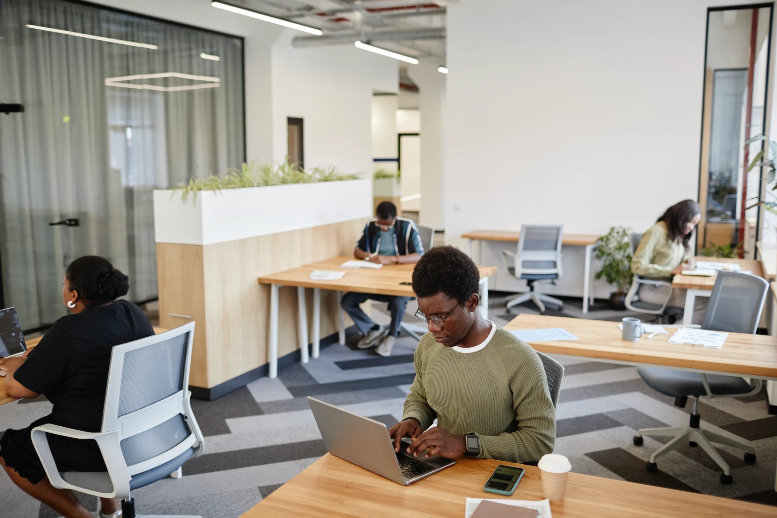 open plan office with multiple people working at wooden tables laptops open and coffee cups nearby